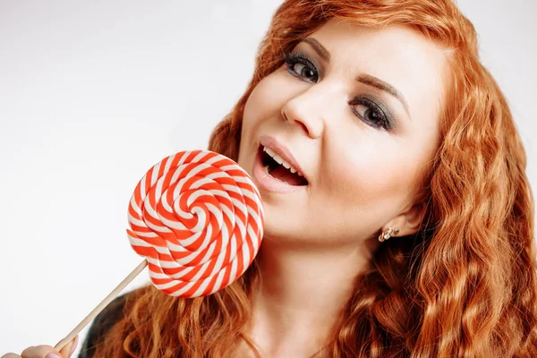 美丽的卷曲红发女人拿着一个红色白色棒棒糖在白色背景 — 图库照片