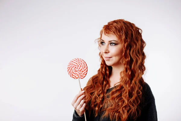 漂亮的年轻的卷曲红头发的女人拿着大甜棒棒糖, 把目光移开 — 图库照片