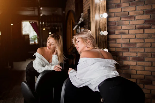 Loira atraente primping no espelho com lâmpadas no estúdio de beleza. Retrato de bela mulher jovem sexy com pele fresca e saudável, maquiagem natural em pé diante do espelho. Salão de cabeleireiro, beleza — Fotografia de Stock