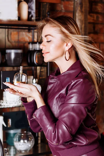 Beauty, aroma, people and body care concept. Young attractive woman with blond flying hair wearing violet leather jacket holding perfume in her hands — Stock Photo, Image