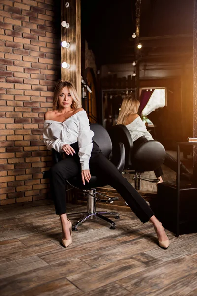 Elegante bella giovane donna sexy in camicia bianca, pantaloni neri seduti sulla sedia accanto al grande specchio. Interni in stile loft. Moda, stile, bellezza, glamour, showroom — Foto Stock
