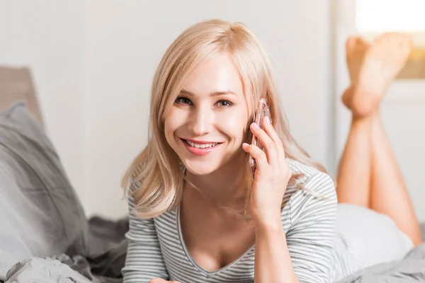 Bonita jovem sorrindo mulher deitada na cama e usando telefone em seu quarto — Fotografia de Stock