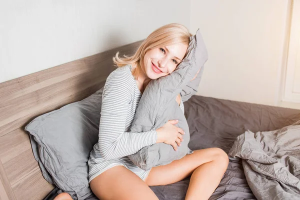 Femme positive avec oreiller assis dans le lit et souriant à la caméra à la maison — Photo