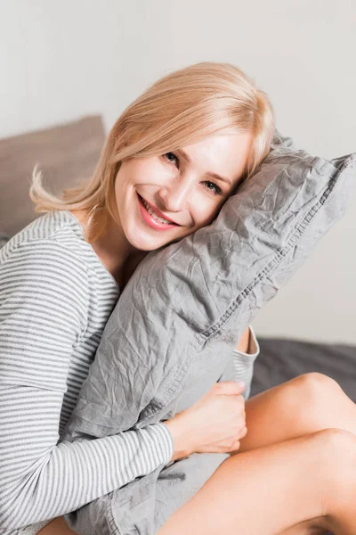Красивая женщина с подушкой сидит в постели и улыбается фотоаппарату дома — стоковое фото