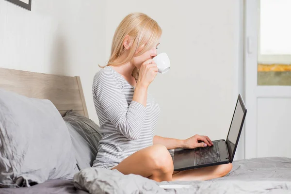 Krásná žena, která používá přenosný počítač a má na posteli šálek kávy — Stock fotografie