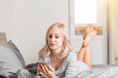 Yatakta yatan ve yatak odasında telefon kullanan güzel genç gülümseyen kadın