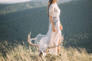 Beautiful girl in long curly white gray dress profile, tulle, veil, nature, mountains, forest, deer skull with horns near the foot, bride, wedding clipart