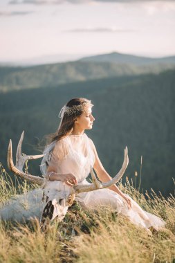 Beautiful girl in a dress sitting with a deer skull with horns on the top of the mountain, eyes closed, decoration on your head, clipart