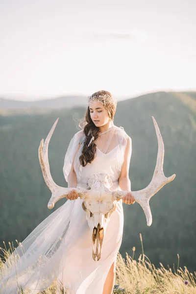 Belle jeune mariée brune en robe longue blanche est faite de tulle regarde vers le bas, garde à portée de main un crâne de cerf avec des cornes décoration sur la tête, la nature , — Photo