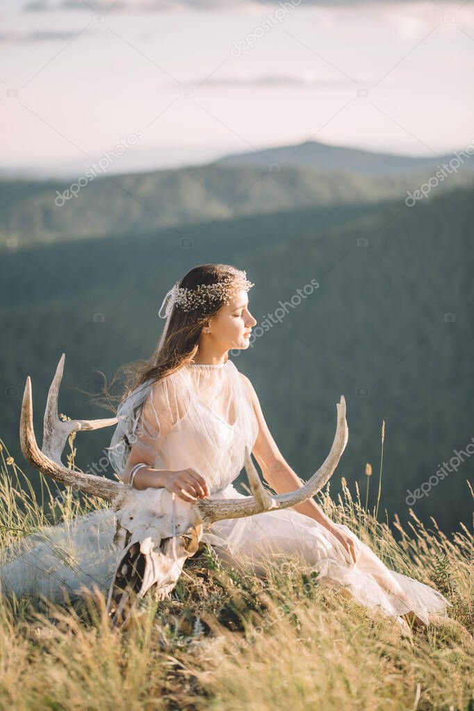 Beautiful girl in a dress sitting with a deer skull with horns on the top of the mountain, eyes closed, decoration on your head,