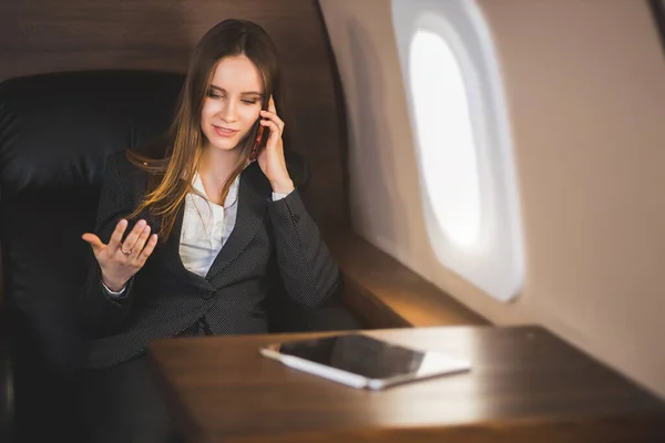 Cheerful happy brown haired business lady in white blouse and formal suit, sitting in comfortable seat, laughing talking on mobile phone during flight on first class plane.
