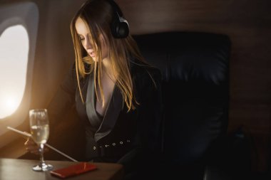 Özel uçakta dijital tablet kullanan çekici bir iş kadını. Beyaz, şık siyah elbiseli hoş bir kadın. Kablosuz kulaklıklı müzik dinliyor ve uçuş sırasında çalışıyor..