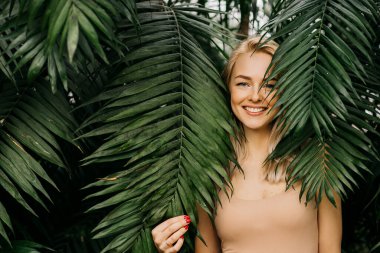 Doğal çıplak makyajlı, mükemmel ciltli, sarı saçlı mutlu bir kadının portresi. Palmiye yapraklarının arkasına saklanan güzel bir kadın. Kozmetik, sağlık, saflık, cilt bakımı, kaplıca konsepti.