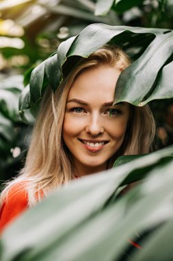 Yağmur ormanlarında poz veren doğal makyajlı sarışın kadın. Kırmızı tişörtlü muhteşem beyaz kadın canavar yapraklarının arasında duruyor, yaz tatilinin tadını çıkarıyor. Vahşi yaşam ve insanlar kavramı.