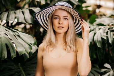 Güneş şapkası takan, ten rengi mayo giyen, mesafeye bakan, tropikal bitki arka planında elini kaldırmış güzel sarışın kadın. Yaz tatili, yüz ifadeleri, duygu konsepti..