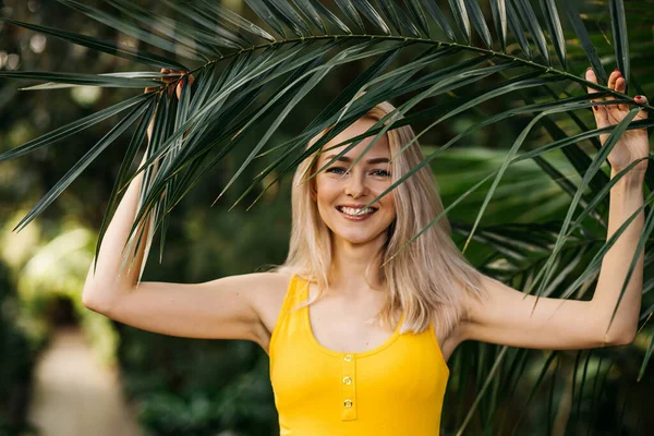Yakından bakınca, mutlu ve gülümseyen genç bir sarışının, egzotik bir ormanda, palmiye ağacının yeşil yapraklarıyla çekilmiş portresi. Yaz, tatil, cilt bakımı konsepti.