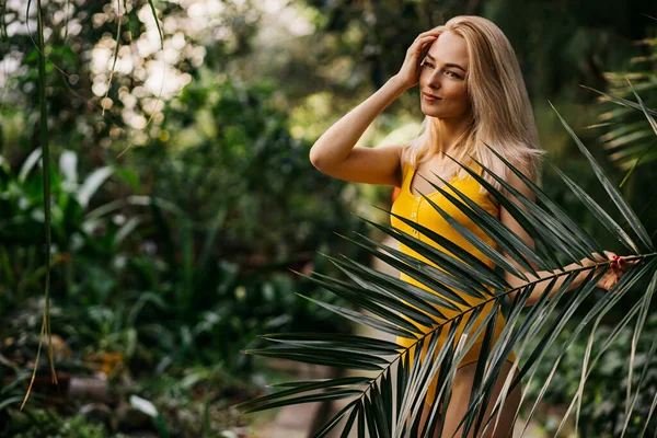 迷人的高加索女人 有着赤裸的妆容和金发 在热带雨林里摆姿势 温柔地触摸着脸 穿着黄色的泳衣 护肤概念 — 图库照片