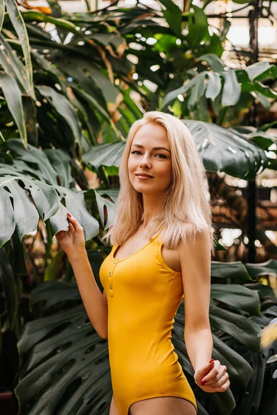 白亜の若い女性の側のビューは エキゾチックな植物の背景に立って 修道院の葉に触れます 黄色のスタイリッシュな水着を着てスリムなブロンドの髪の女性のプロフィールショット — ストック写真