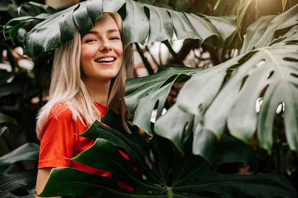 Kırmızı tişört giyen neşeli, beyaz dişli, beyaz dişli, tropikal yaprakların arasında dikilen beyaz tişörtlü, beyaz tenli sarışın kadın. Doğa ve insan kavramı.