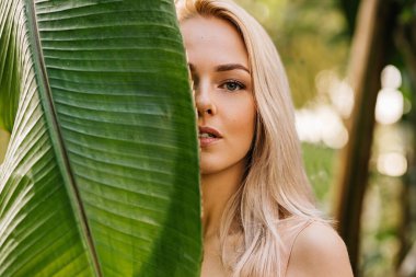 Sarışın, doğal makyajlı, temiz tenli, yüzünün yarısını tropikal yaprağın arkasına saklayan beyaz bir kadının yakın plan portresi. Cilt bakımı, kaplıca, güzellik, organik kozmetik ürünler konsepti.