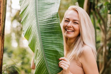 Attractive lady with natural beauty posing in rainforest. Sensual gorgeous blonde woman in flesh color swimsuit standing near big tropical tree, enjoying summer vacation. Nature and people concept. clipart
