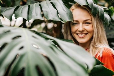 Yağmur ormanlarında poz veren doğal makyajlı sarışın kadın. Kırmızı tişörtlü muhteşem beyaz kadın canavar yapraklarının arasında duruyor, yaz tatilinin tadını çıkarıyor. Vahşi yaşam ve insanlar kavramı.