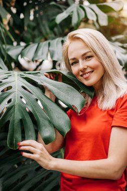 Yağmur ormanlarında poz veren doğal makyajlı sarışın kadın. Kırmızı tişörtlü muhteşem beyaz kadın canavar yapraklarının arasında duruyor, yaz tatilinin tadını çıkarıyor. Vahşi yaşam ve insanlar kavramı.