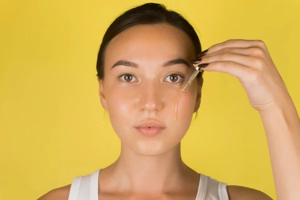 Uma jovem linda aplica um soro no rosto com uma pipeta. Cosmetologia, cuidados com a pele. Fundo amarelo — Fotografia de Stock