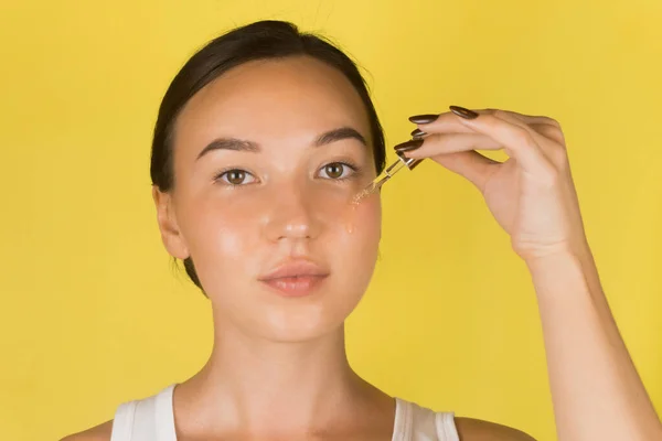 Seorang gadis muda yang cantik mengaplikasikan serum ke wajahnya dengan pipet. Kosmetologi, perawatan kulit. Latar belakang kuning — Stok Foto