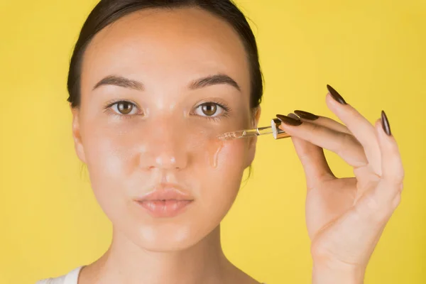 Güzel bir genç kız yüzüne pipetle serum uyguluyor. Kozmetoloji, cilt bakımı. Sarı arkaplan