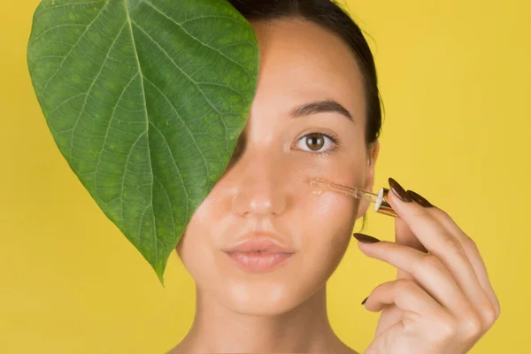 Seorang gadis muda yang cantik mengaplikasikan serum ke wajahnya dengan pipet. Kosmetologi, perawatan kulit. Latar belakang kuning — Stok Foto