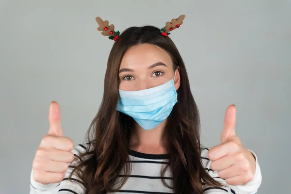 Bella giovane bruna con una maschera protettiva medica su uno sfondo grigio. Una donna posa con una decorazione natalizia tra i capelli. Festeggiamo il nuovo anno nel mezzo di una pandemia. — Foto Stock