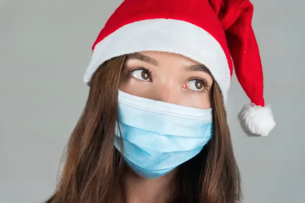 一个戴着圣诞老人帽和医疗面具的年轻女子看着摄像机 在大流行病中庆祝新年 是的高质量的照片 — 图库照片