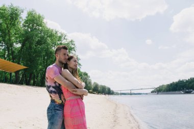 Genç çift, sarılma ve nehir kıyısında birbirlerine gülümsüyor. Kumlu sahilde yürüyüş