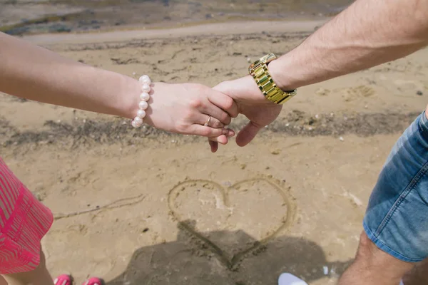 A young couple holds hands by the river. In the sand, a heart is drawn