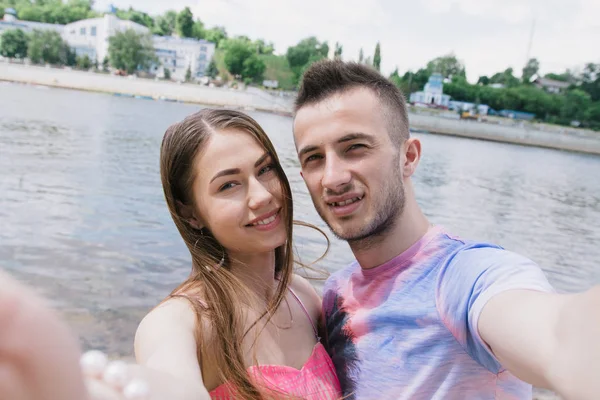 Νεαρό ζευγάρι αγκαλιάζει και να χαμογελούν σε κάθε άλλο στις όχθες του ποταμού. Περπατώντας κατά μήκος της αμμώδους παραλίας. Βεβαιωθείτε Selfies — Φωτογραφία Αρχείου