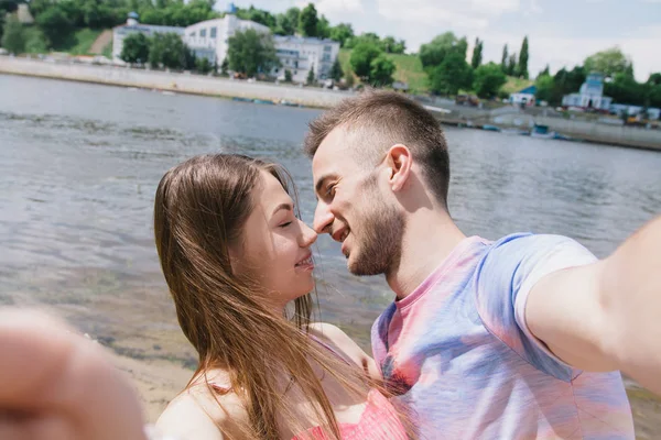 Jong (echt) paar knuffelen en glimlachend bij elkaar op de rivieroever. Een wandeling langs het zandstrand. Selfies maken — Stockfoto