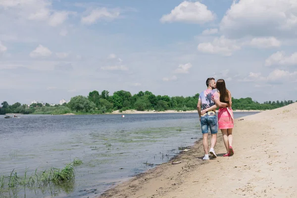 Jong (echt) paar knuffelen en glimlachen naar elkaar op de rivieroever. Wandelen langs het zandstrand — Stockfoto