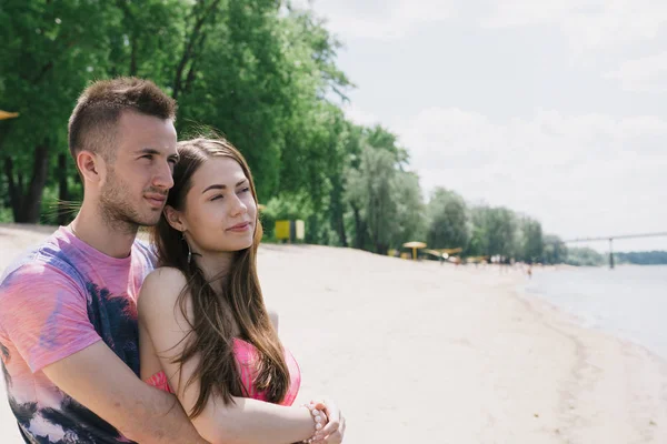 Casal jovem abraçando e sorrindo um para o outro na margem do rio. Caminhando ao longo da praia de areia — Fotografia de Stock