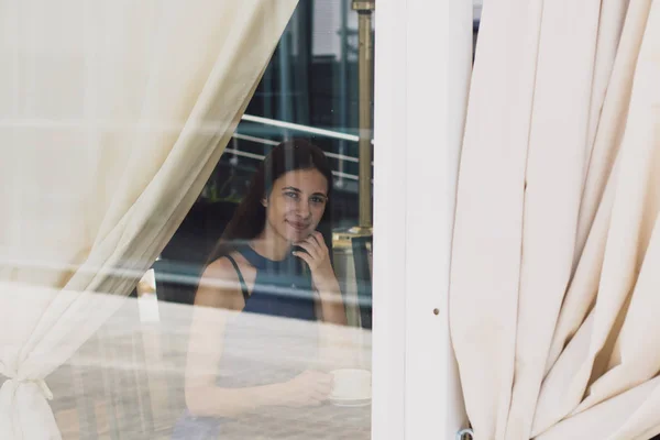 Очень красивая деловая женщина сидит в кафе и держит чашку кофе в руках за стеклом — стоковое фото