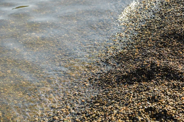 Guijarros en la playa guijarros en la orilla del mar y olas de agua iluminadas por los rayos del sol . —  Fotos de Stock