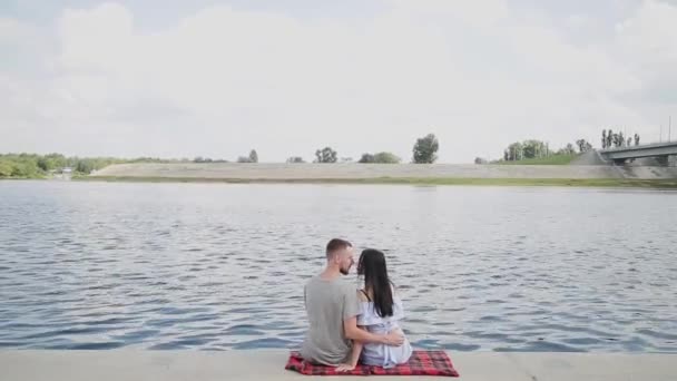 Una pareja amorosa sentada junto al río besándose y abrazándose . — Vídeo de stock