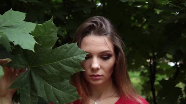Dívka v červených šatech s velkým listem zelené maple na její tváři. — Stock video