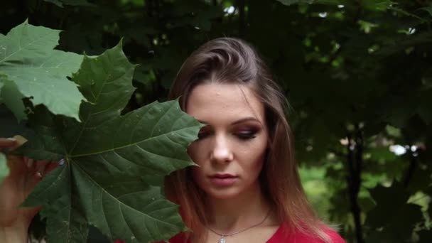 Una chica en un vestido rojo con una hoja grande de un arce verde en su cara . — Vídeos de Stock