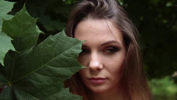 Una chica en un vestido rojo con una hoja grande de un arce verde en su cara . — Vídeos de Stock