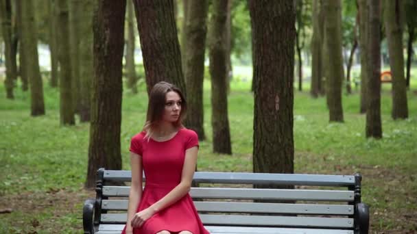 Jeune belle fille dans une robe rouge est assis sur un banc dans le parc . — Video