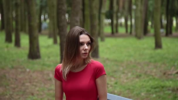 Mooi meisje in een rode jurk zit op een bankje in het park. — Stockvideo