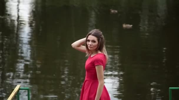 Μια πολύ όμορφη κοπέλα σε ένα κόκκινο φόρεμα στέκεται σε μια προβλήτα κοντά σε μια μικρή λίμνη. — Αρχείο Βίντεο