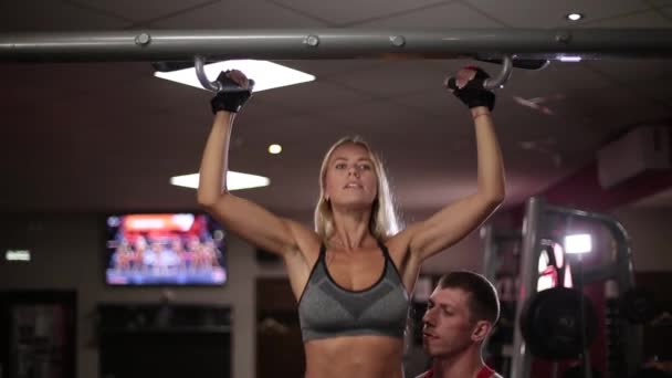 Une femme dans la salle de gym se serre sur la barre transversale avec l'aide d'un instructeur . — Video