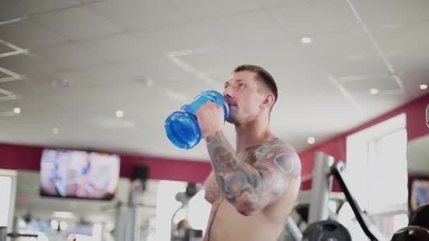 Un hombre está bebiendo agua en el gimnasio con una botella azul después del entrenamiento . — Vídeo de stock